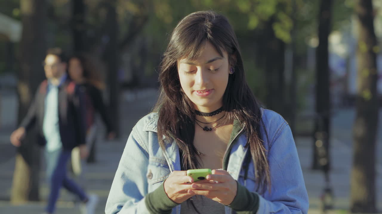Ragazza adolescente sorridente che usa lo smartphone mentre passeggia in un viale del parco.