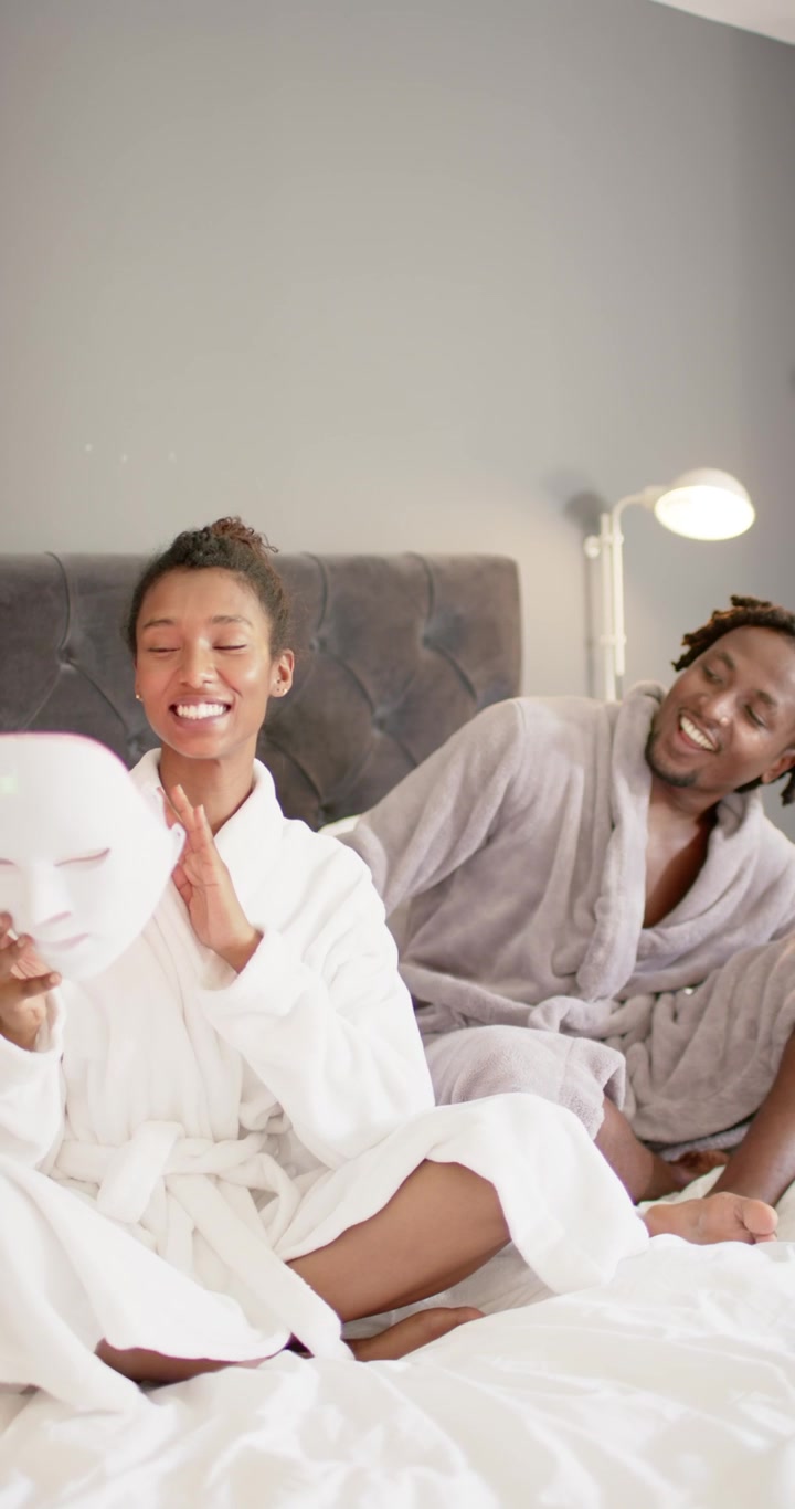 Vertikal video: Afroamerikansk par sitter på sengen i morgenkåper, kvinne holder maske, mann fikser stropp.