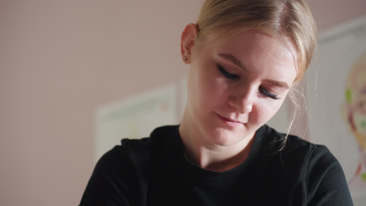 Close-up of light skin woman with blonde hair smiling gently while looking down, wearing black outfit in softly lit indoor setting with calm atmosphere, showing relaxed facial expression
