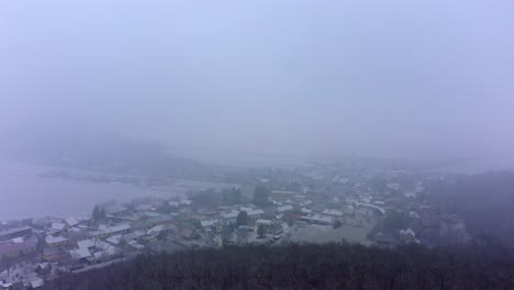 Drone-flies-across-the-moody-forest,-approaching-the-snowy-village-nearby