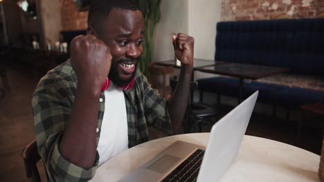 a man with a beard in red headphones and a plaid shirt works on a laptop in a cafe, rejoices in success and clasps his hands