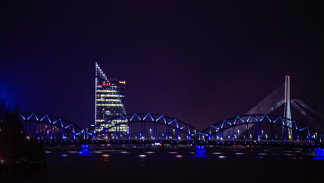 Puente-De-Arco-Del-Ferrocarril-De-Riga-Sobre-El-Río-Daugava-Con-Puente-Vansu-Y-Sede-De-Swedbank-En-El-Fondo-Por-La-Noche-En-Letonia