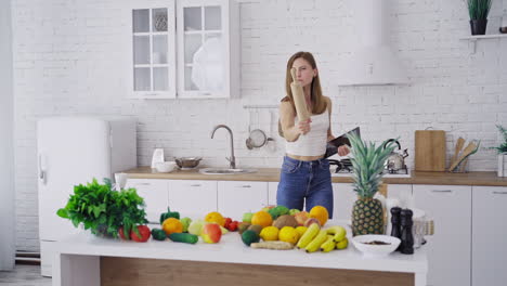 Beautiful woman with the rolling pin in the kitchen. Young housewife shows angry face with kitchen tools. Fresh organic fruit and vegetables on the table