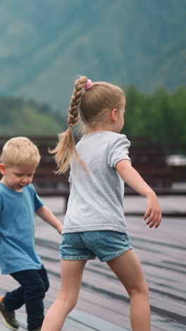 little girl with plaited ponytail and funny younger brother run along wooden dance deck against mountain silhouettes on spring day slow motion