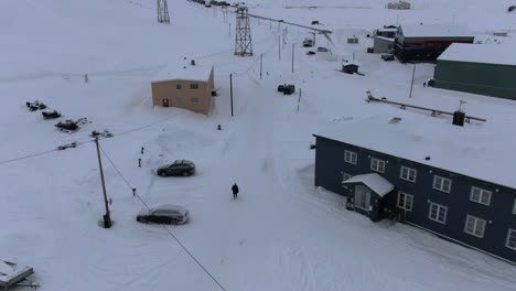 drone view in svalbard flying over longyearbyen town showing a person in a black coat walking between houses in a snowy area with mountains in norway