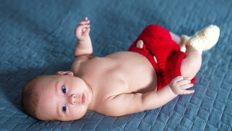 blue-eyed caucasian baby boy lying on bed wearing red shorts. cute kid with naked belly tossing feet. grey backdrop.