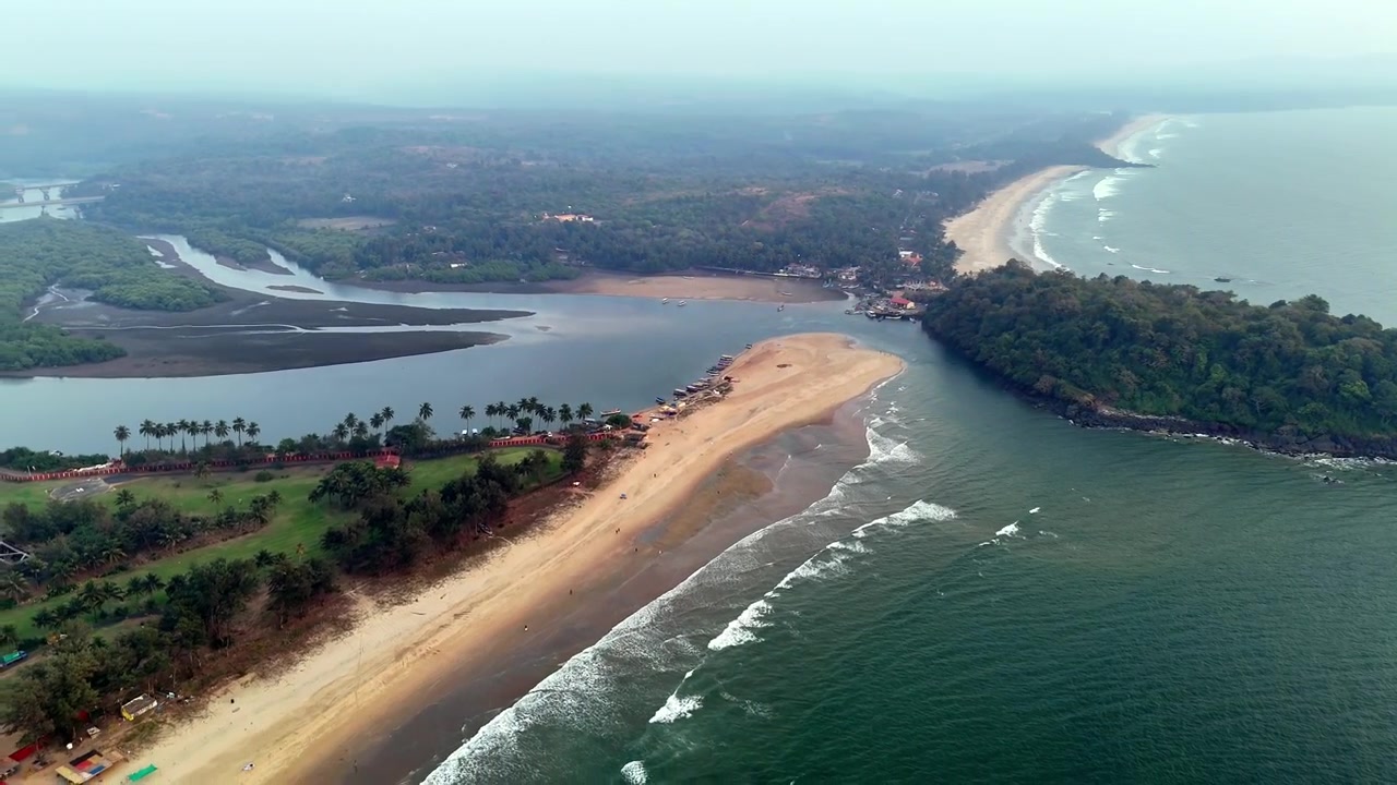 Filmový letecký pohled na The LaLiT Golf and Spa Resort Goa, zasazený mezi bujnou zelení, říčními rameny a Arabským mořem, s vlnami valícími se podél pobřeží pod měkkou, mlhavou oblohou.
