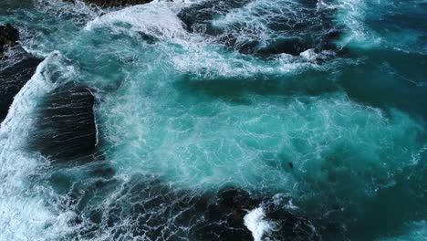 Aerial-drone-shot-of-waves-crashing-up-against-rocks-on-a-shoreline