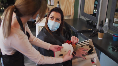 peluquería femenina con máscara facial mostrando una tabla de muestras de color de cabello a una cliente femenina