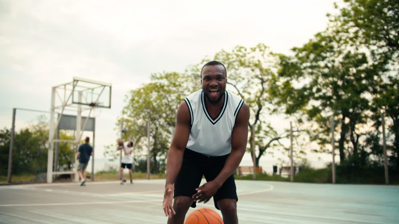 A young Black man in a white t-shirt shows how he skillfully handles a basketball on a basketball court outside in summer