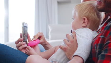 Young-white-couple-at-home-with-their-baby-using-smartphone