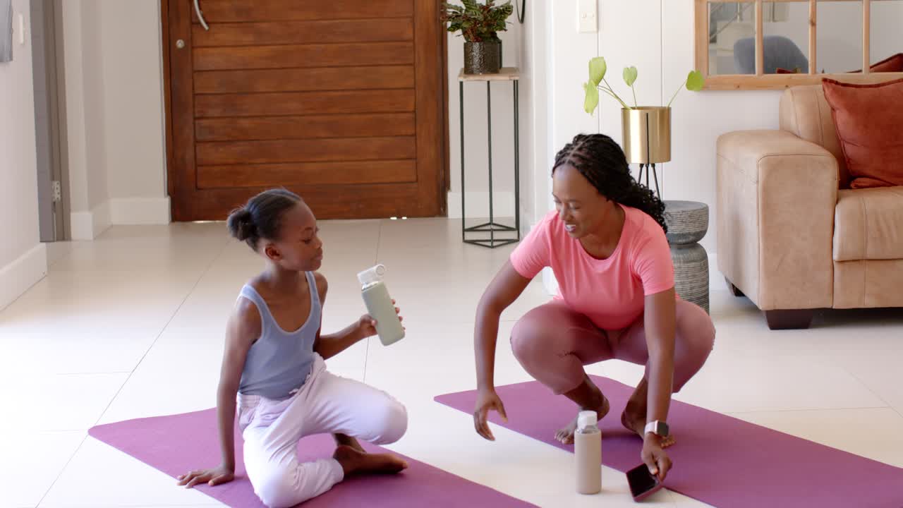 African American mom and daughter entering at home holding bottles placing phone on mats stretching