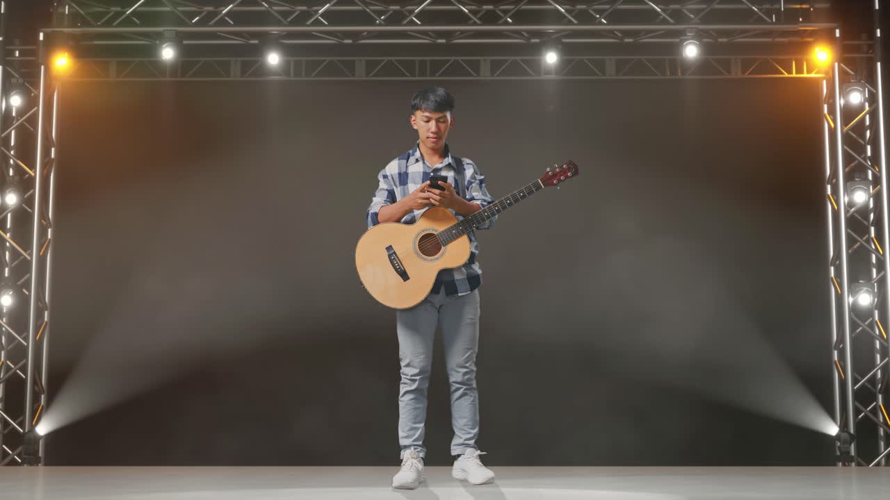 cuerpo lleno de adolescente asiático con guitarra apuntando a teléfono inteligente en la mano en el escenario