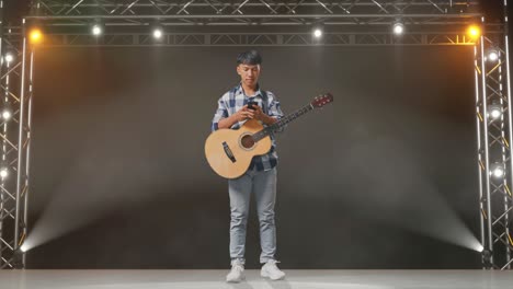 full body of asian teenager with guitar pointing to smartphone in hand on stage