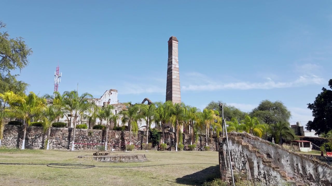 Vista a nivel del suelo de la histórica chimenea en Hacienda Coahuixtla rodeada de palmeras, muros de piedra y ruinas conservadas bajo un cielo azul claro en Morelos, México