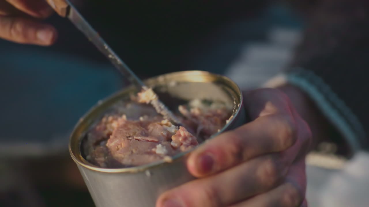 close up of person in green sweater kneeling on forest floor scooping meat from open metal can using knife while seated on ground during rustic camping meal near glowing tent under soft natural light