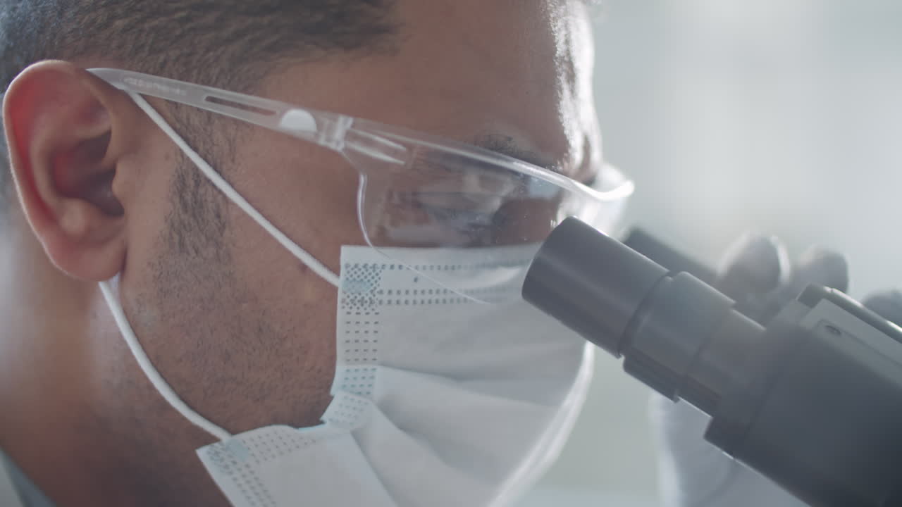 Scientist in Mask, Glasses and Gloves Using Microscope