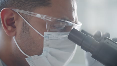 científico con máscara, gafas y guantes usando el microscopio