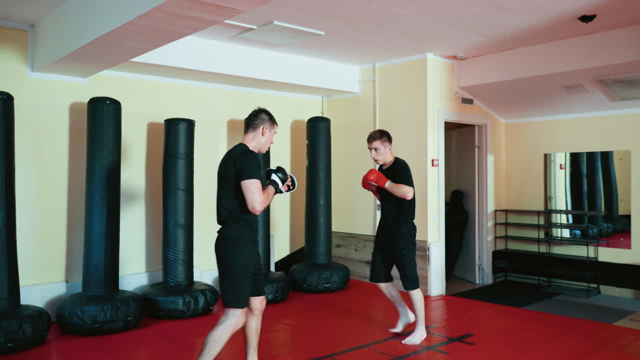 Fighters sparring on red mat inside martial arts gym, wearing gloves and black outfits, facing each other in close stance to exchange strikes during intense training surrounded by punching bags indoors