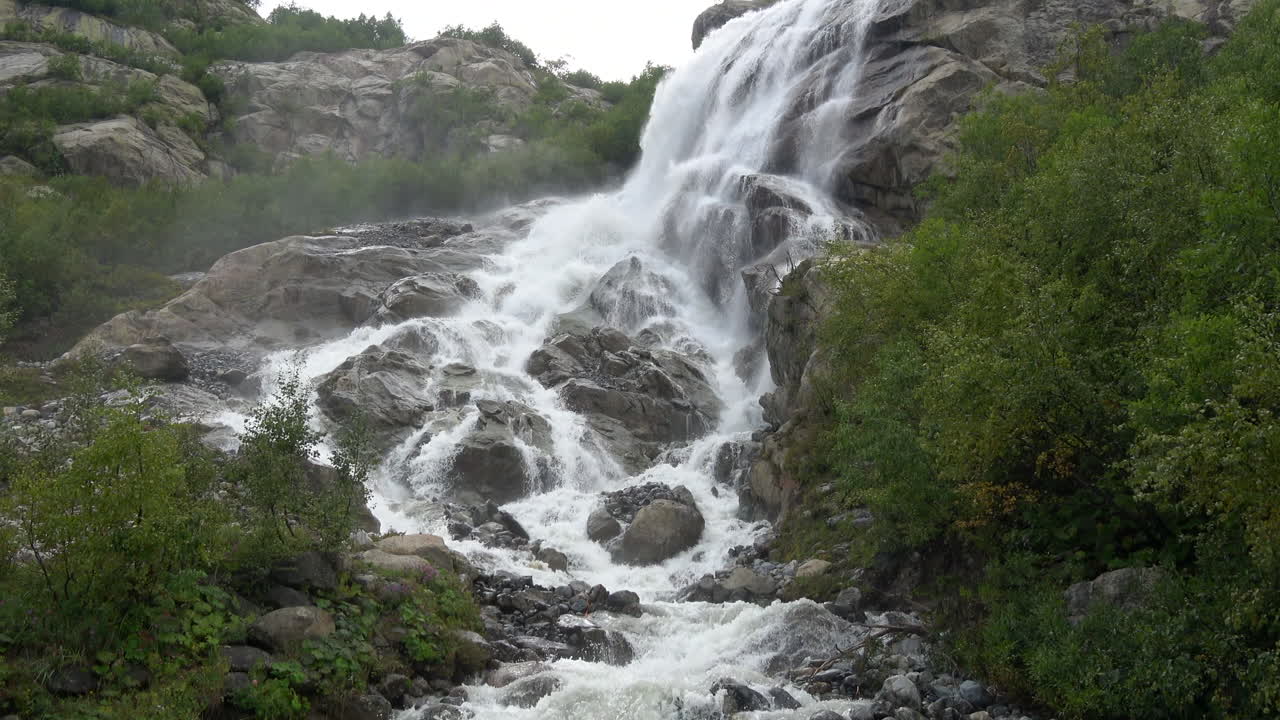 Cascata scenografica che si getta impetuosa su rocce scure in un ruscello di montagna, circondata da verdi pendii boscosi a Dombay, Caucaso, Russia, ideale per temi di viaggio e natura.