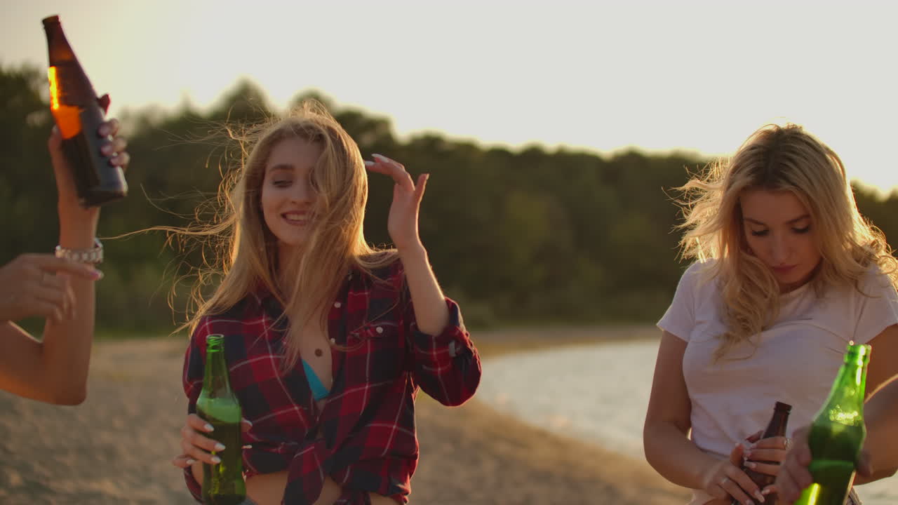 Giovani donne festeggiano un compleanno a una festa all'aperto con amici, birra e buon umore. Ballano e i loro capelli volano in una sera d'estate vicino alla riva del fiume