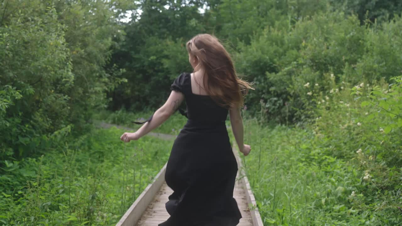 Woman in black dress walking on a wooden boardwalk in a lush green park