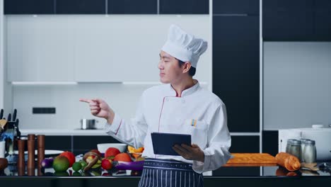 asian man chef using a tablet and pointing to side while standing in home kitchen