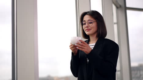 woman in black jacket stands at the window drinking coffee. smiling lady looks at window enjoying her drink. low angle view.