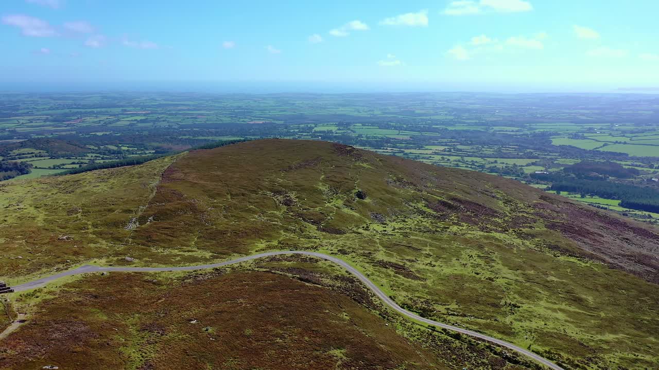 Luftfoto af vej, der krydser en bakketop med en skov og udsigt i det fjerne i baggrunden om sommeren i det sydlige Irland.