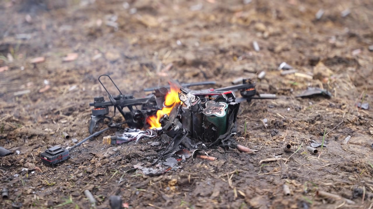 Drone FPV che cova su terreno sterrato con fumo e segni di bruciatura in un ambiente esterno.