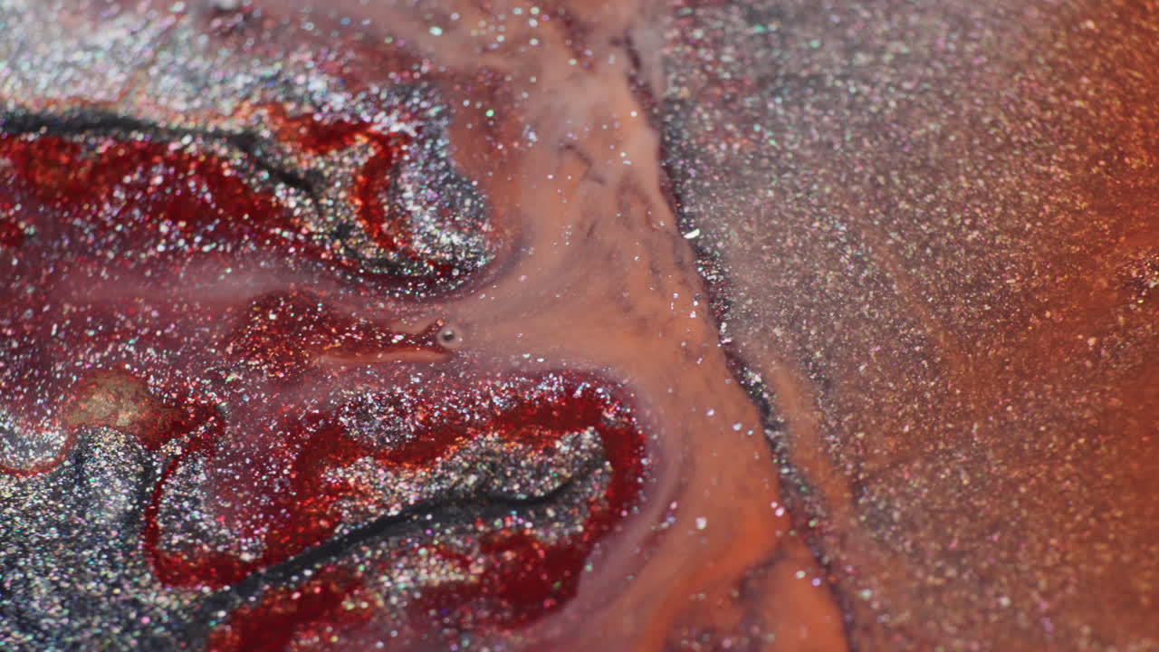 Macro shot showing glittery grey and orange ink reacting to added water. The red ink spreads and mixes with the grey, forming mesmerizing, fluid patterns