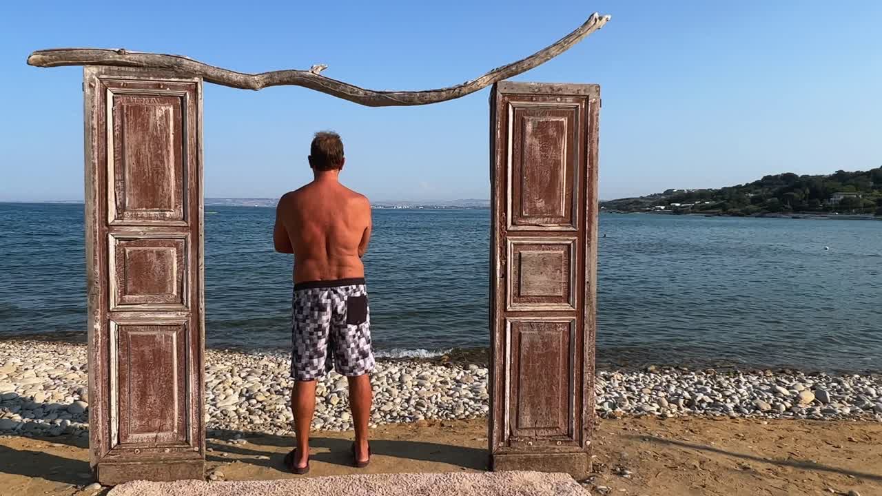 Deniz kenarında dekoratif ahşap kapı ve mayo ile ayakta duran, kapı pervazına yaslanmış bir adamın arkadan görünüşü.