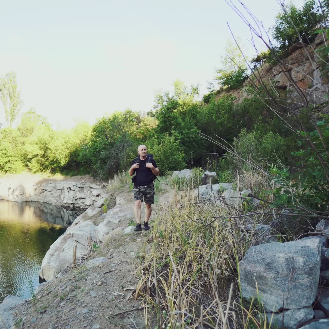 traveler is holding hands for the harnesses of a backpack and walking along the bank on the background of stones and rock. The tourist is looking at the wonderful landscape around him