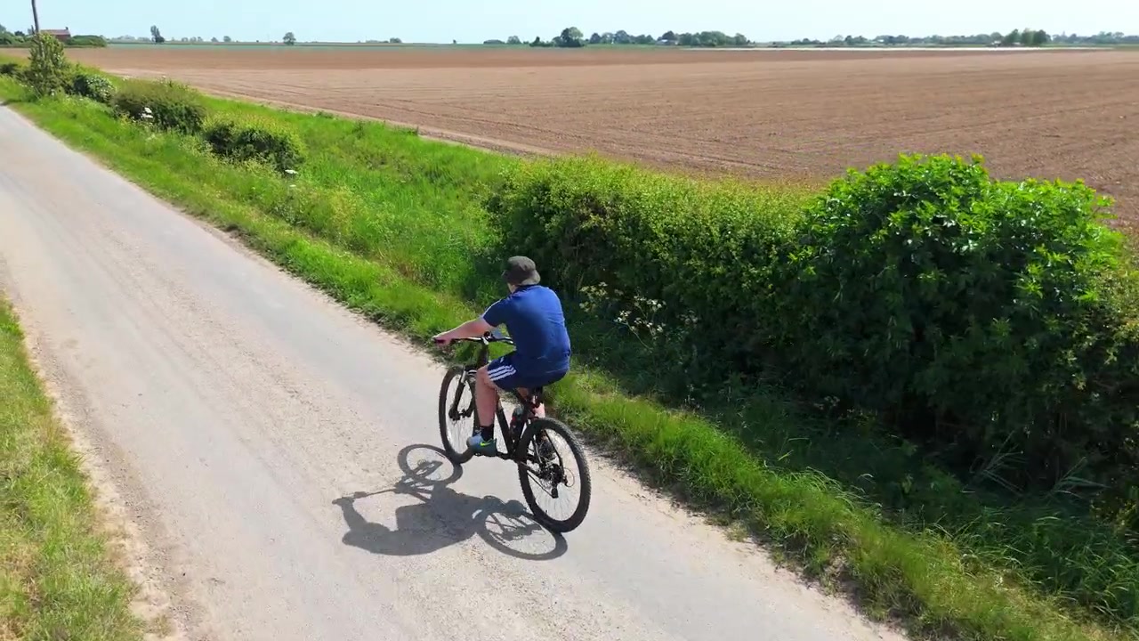 Luftfoto af en person, der cykler på en smal landevej. Grønne hække flankerer stien, med marker af afgrøder, der strækker sig ud i det fjerne. Cyklisten virker rolig og tilfreds.