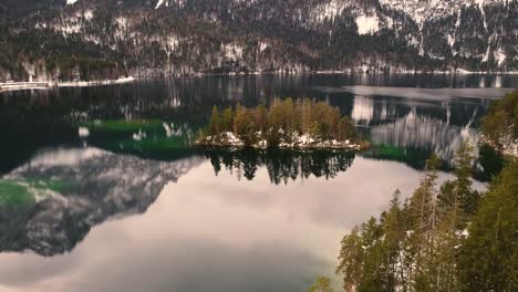 aerial view of tiny island covered with pine trees in middle of clear lake reflecting surrounding snowy mountains and sky, creating peaceful and cinematic winter landscape in iceland
