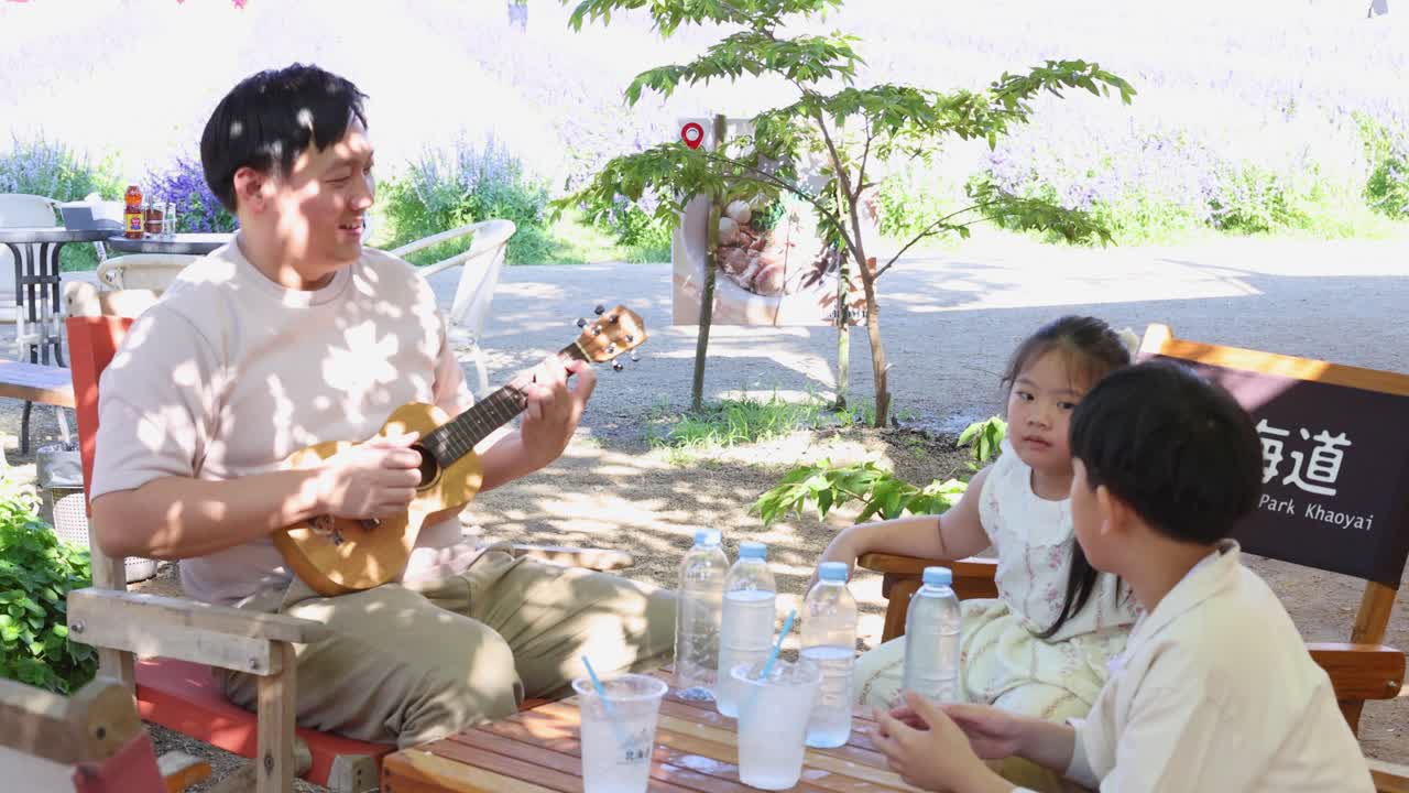 คุณพ่อมีความสุขเล่นอูคูเลเล่ให้ลูก ๆ สองคนฟังที่คาเฟ่กลางแจ้งที่มีแดดส่อง บรรยากาศครอบครัวที่ผ่อนคลาย