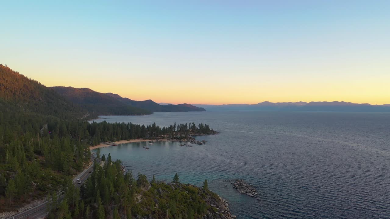 Cars Driving by Beautiful Lake Taho with a Colorful Sunset and a Pine Tree Forest with Clear Blue Water and Mountains in the Background - aerial drone reveal