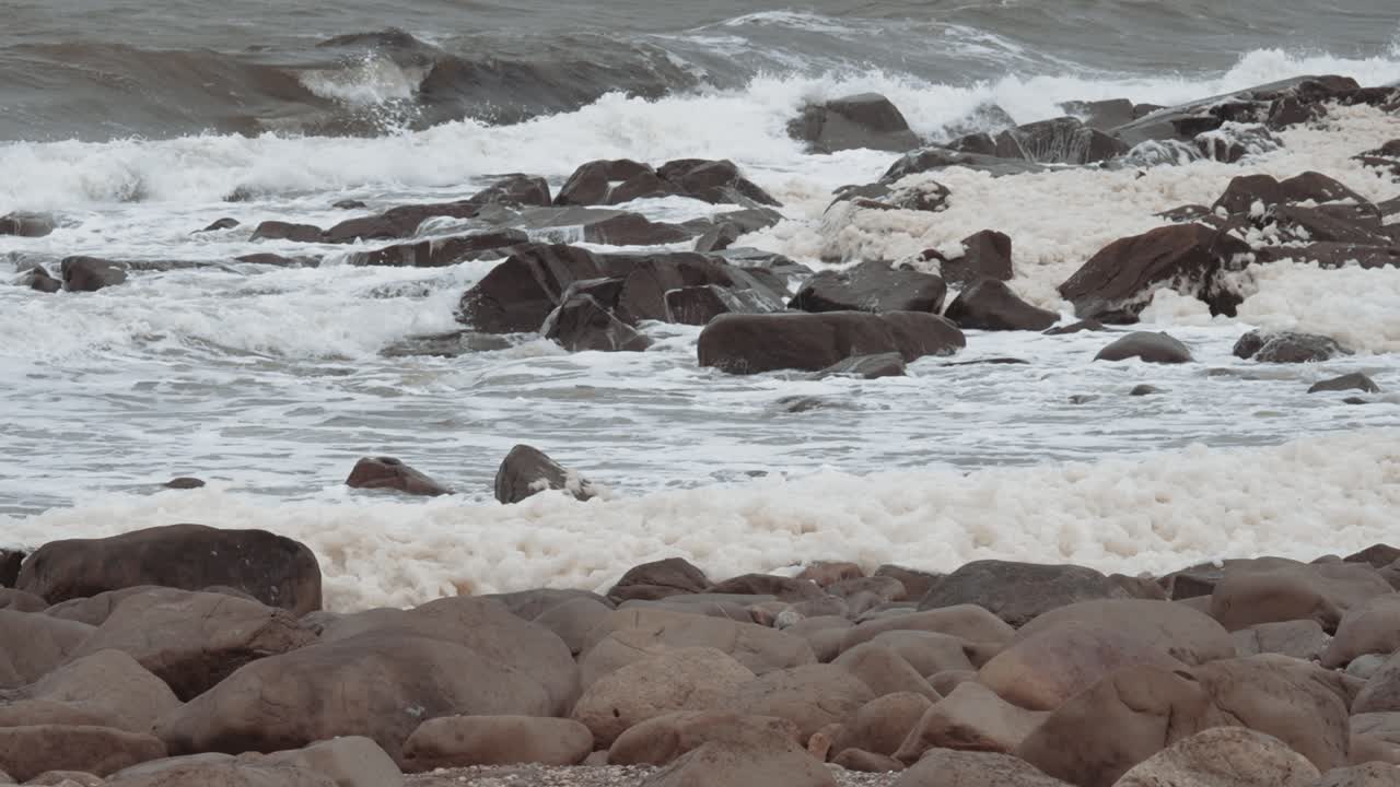 천천히 움직이는 바다 거품이 바위 모양의 해변을 가로질러 불고 있으며, 배경에는 파도가 흐르고 있습니다.