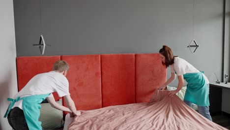 View-from-the-side-of-a-confident-cleaning-guy-a-blond-guy-with-stubble-and-in-a-white-T-shirt-with-a-blue-front-together-with-his-colleague-a-brunette-girl-making-the-bed-in-a-modern-apartment-while-cleaning-on-call