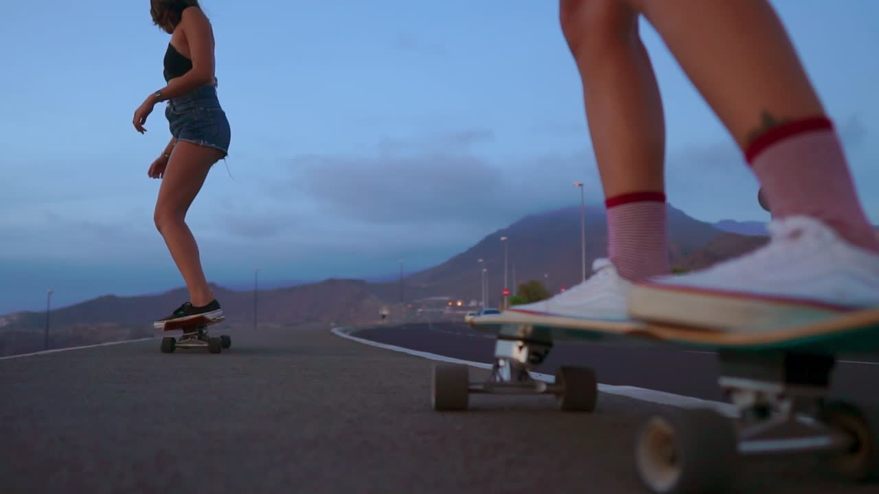 In slow motion, two friends skateboard on a road at sunset, with mountains and a beautiful sky enhancing the scenery. They're dressed in shorts