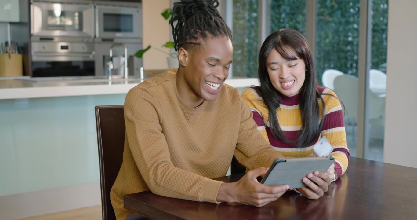 Diverse couple in sweaters at kitchen table, tablet displaying, tapping, sharing card, reviewing