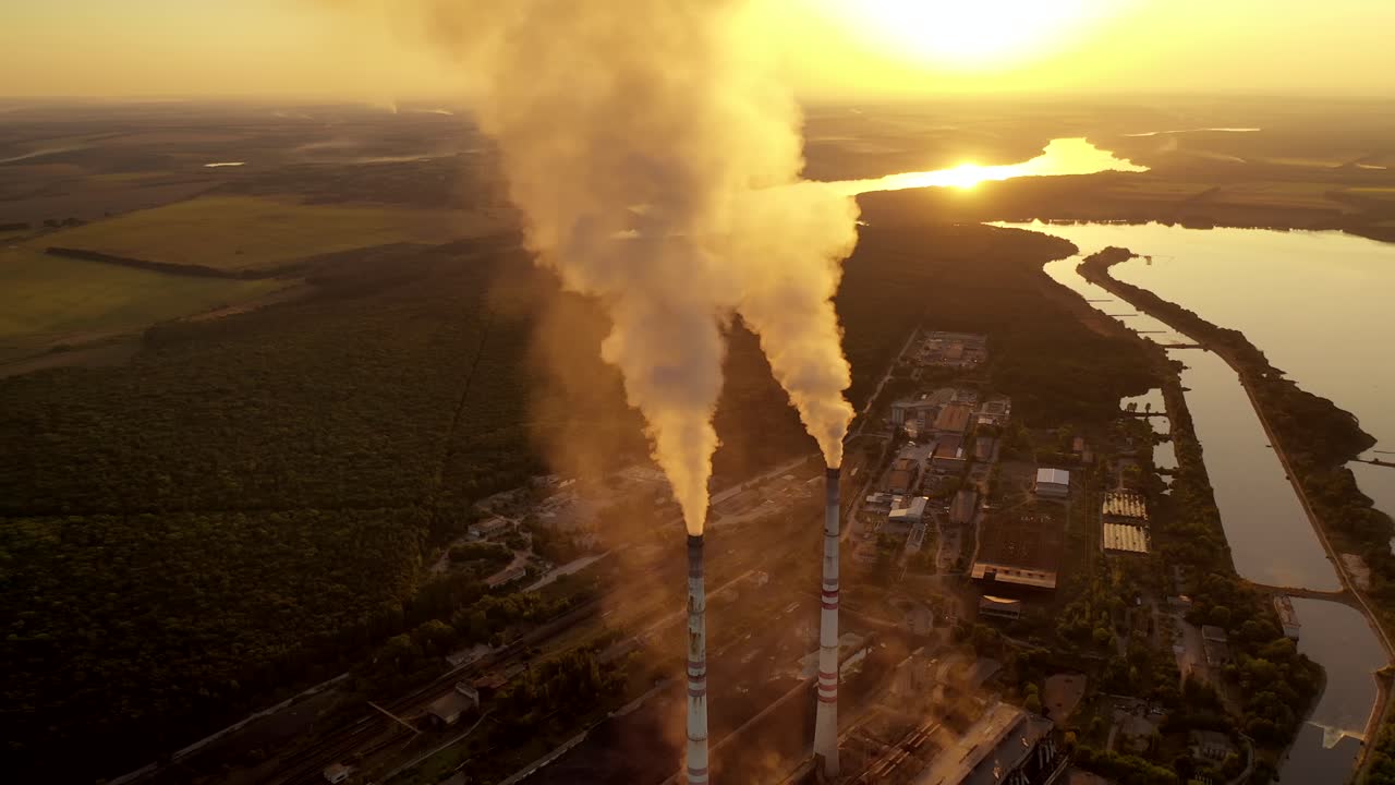 Manufacturing in nature at sunset. White smoke goes from metallurgical pipes into the air. Industrial factory near the river among fields. Aerial view.