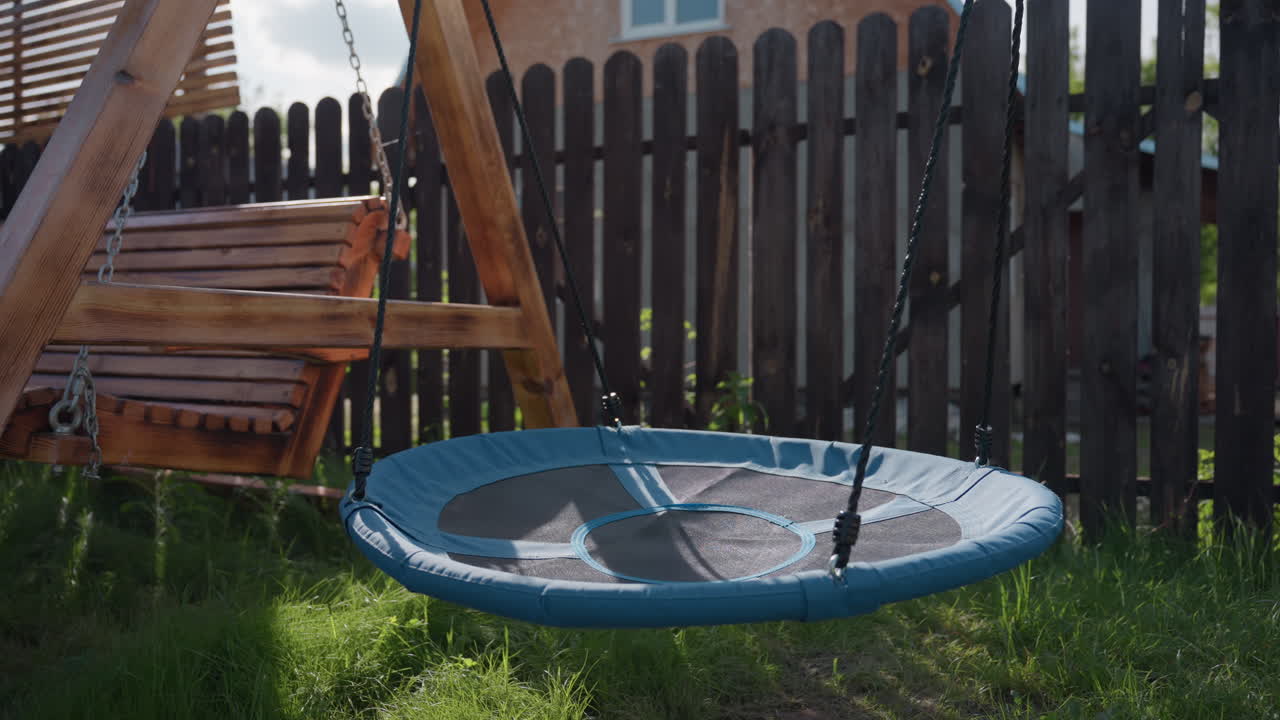 Close up of backyard playground featuring wooden bench swing and circular blue nest swing suspended above soft grass, sunlight filtering through slatted panels beside dark wooden fence