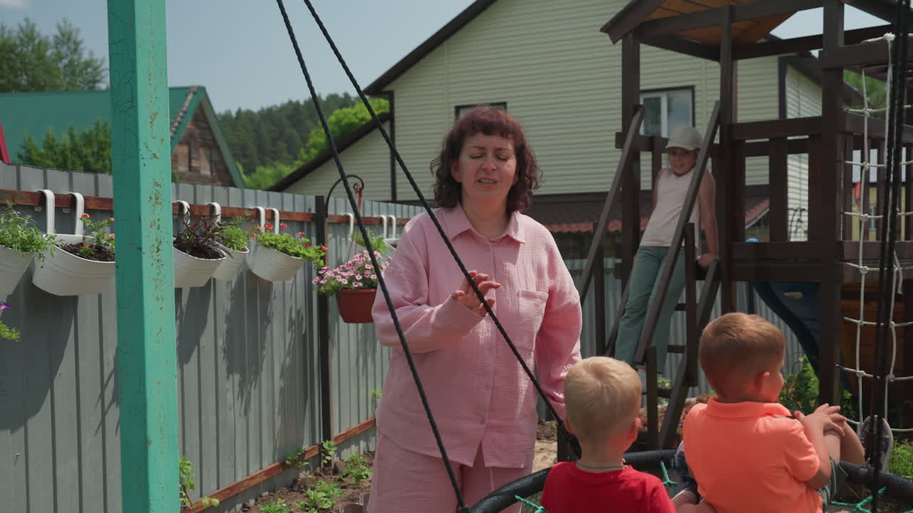 Woman Watches Kids Playing, Adult Woman Attentively Observing Children At Playground During Summer, Caucasian Woman Carefully Supervising Children At Outdoor Playground On Summer Day