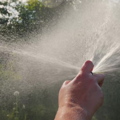 Close-up-of-hand-with-garden-hose-watering-garden-and-trees-in-garden