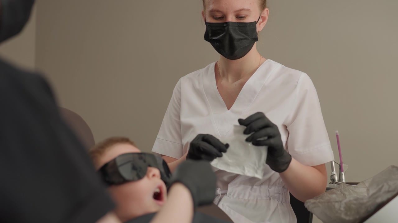 Nærbillede af tandlægeassistent der forbereder materialer til ortodontisk procedure med ung patient iført beskyttelsesbriller. Tandlæge i hvid uniform og sorte handsker håndterer gaze