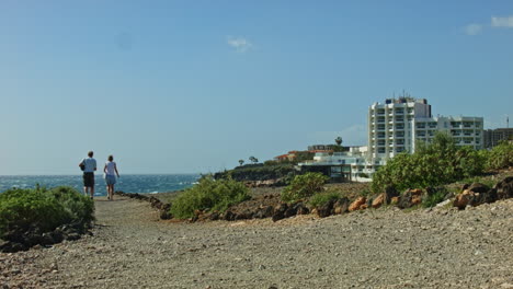 Tenerife-Beach-Road-Turistas-Caminando-Con-Un-Hotel-Y-Olas-En-El-Fondo-Y-Sendero-Rocoso,-Carretera