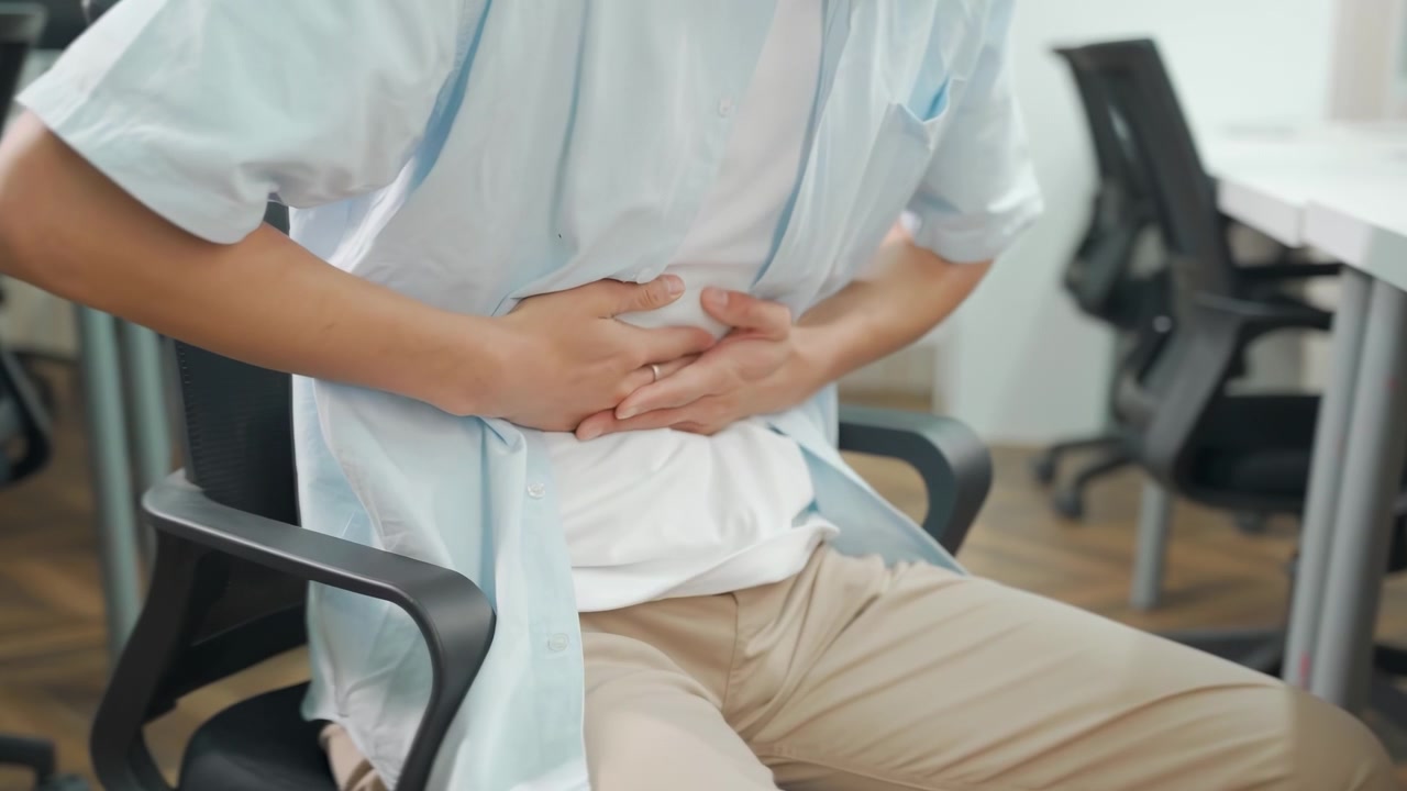 Young businessman experiencing typing on laptop in office and suffering stomach pain. Concentrated male in formal wear holding belly and experiencing acute attack of pain in abdomen during work