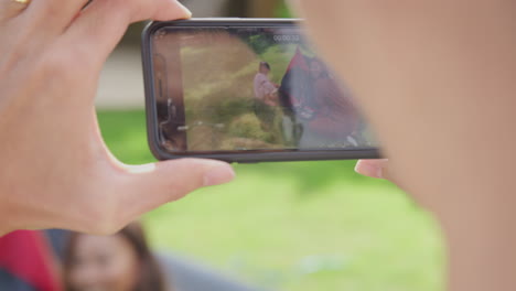 Primer-Plano-De-Un-Padre-Asiático-Filmando-Un-Vídeo-En-Un-Teléfono-Móvil-Mientras-La-Familia-Montaba-Una-Tienda-De-Campaña-En-El-Jardín-De-Casa