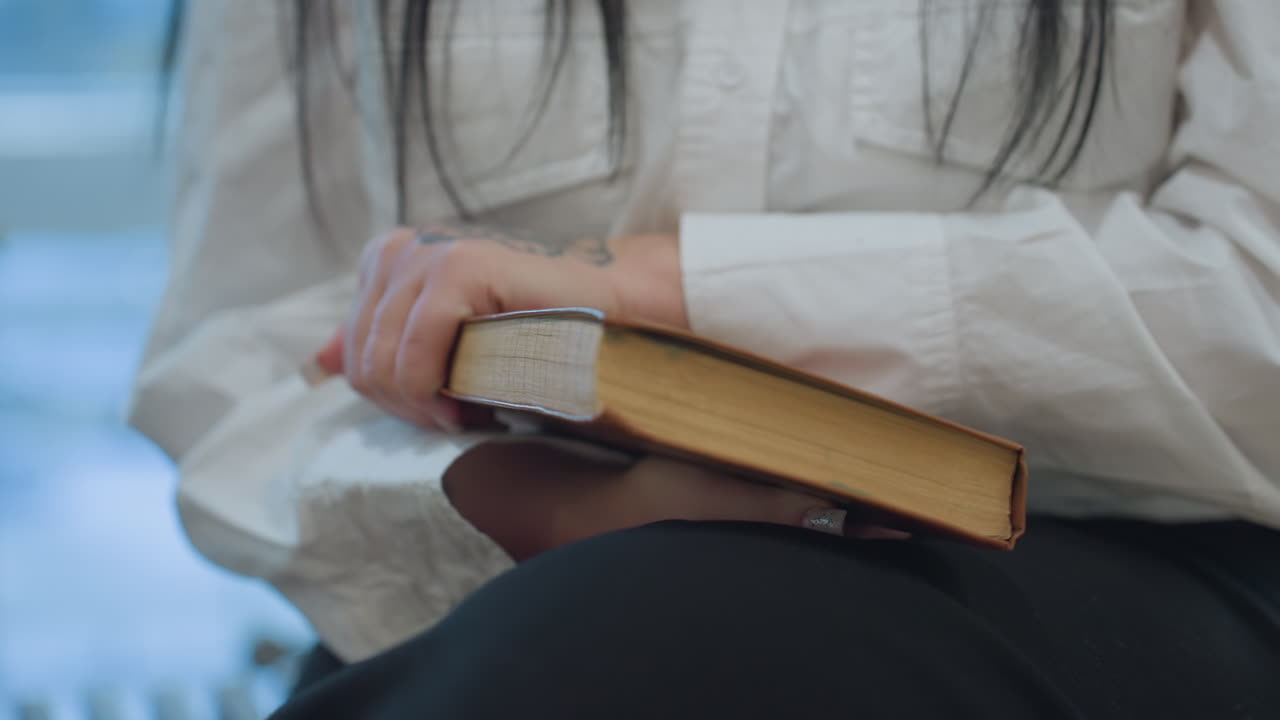 Close up on woman manicured nails gripping hardcover book then slamming it shut in frustration, stray pages trembling, blurred modern interior background